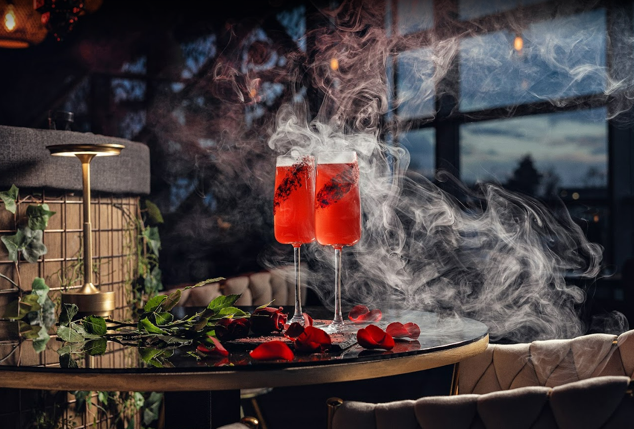 Two coupe cocktails with rose petals and dramatic smoke on a candlelit table